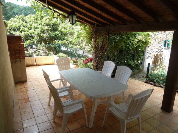 Un patio al aire libre con mesa y sillas, rodeado de follaje y un muro de piedra.