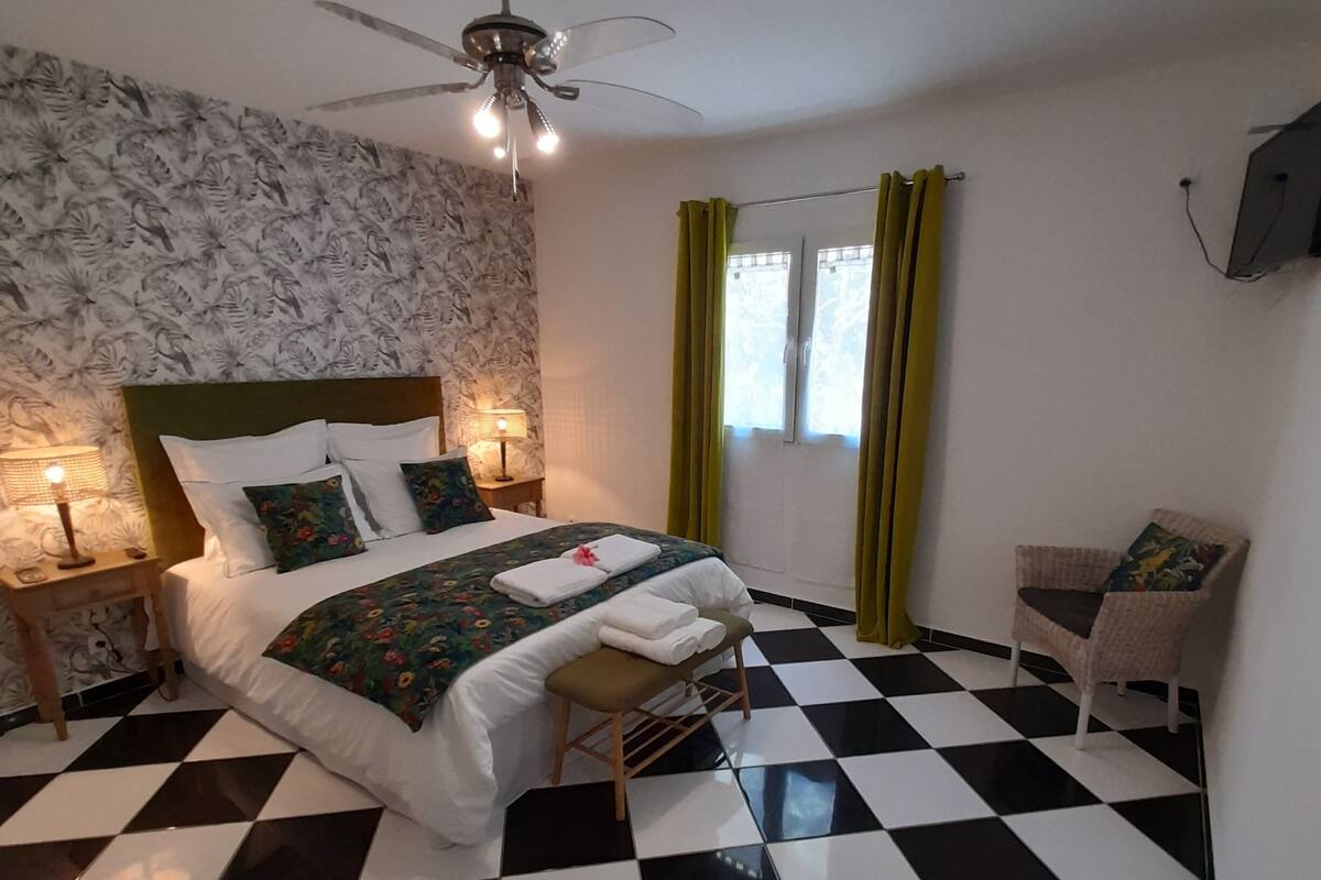 A hotel room with a checkered floor, a bed with white linens, a ceiling fan, a nightstand with a lamp, and a window with curtains.