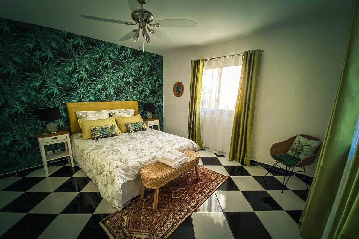 A bedroom with a checkered floor, a bed with a patterned bedspread, a ceiling fan, and a window with curtains.