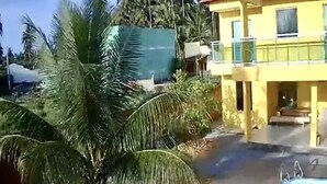 Un edificio amarillo con balcón, rodeado de palmeras y una piscina.