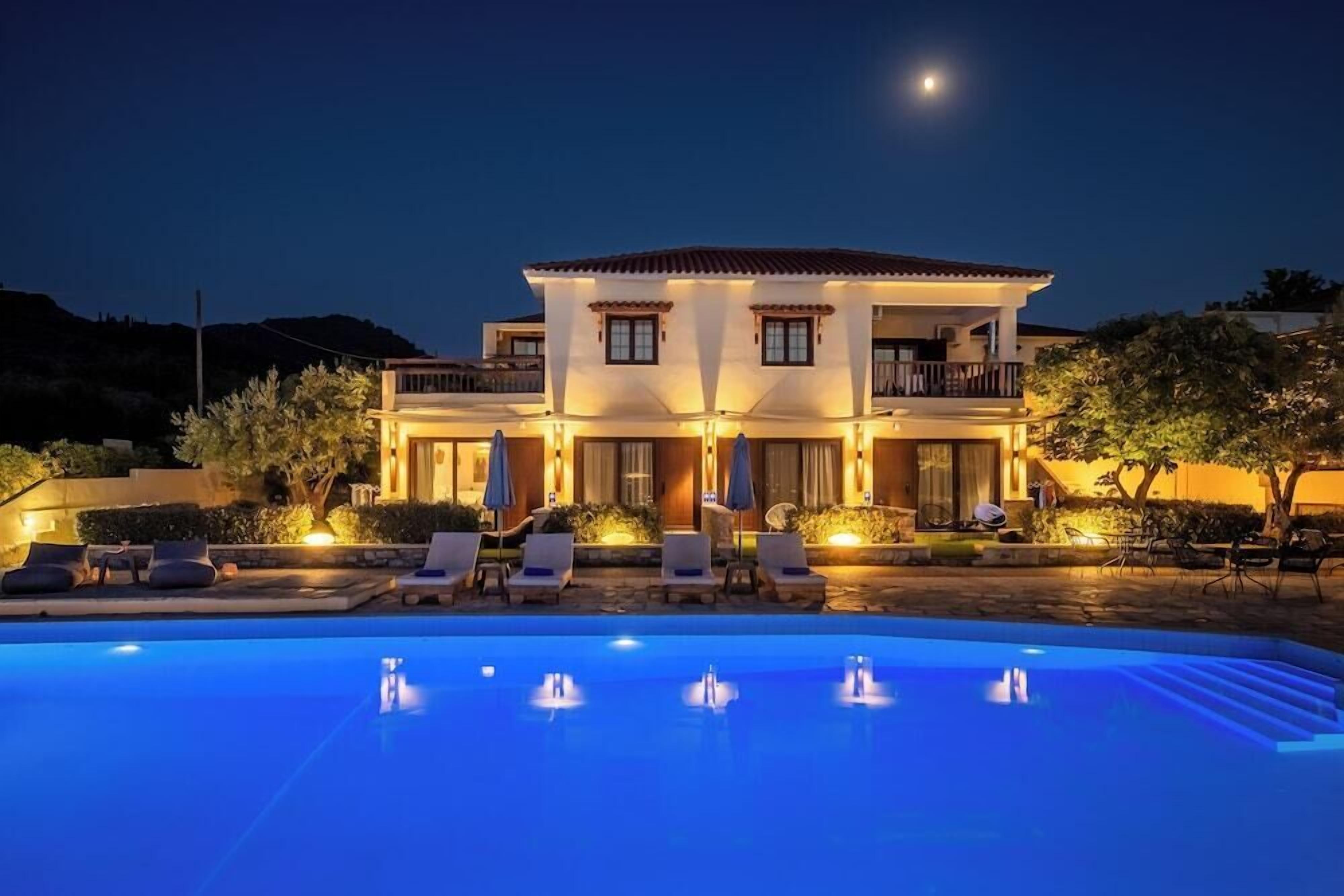 A two-story house with a swimming pool in front, surrounded by lounge chairs and umbrellas.