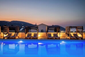 Un área junto a la piscina con sillas de descanso y sombrillas, cielo despejado y colinas al fondo.