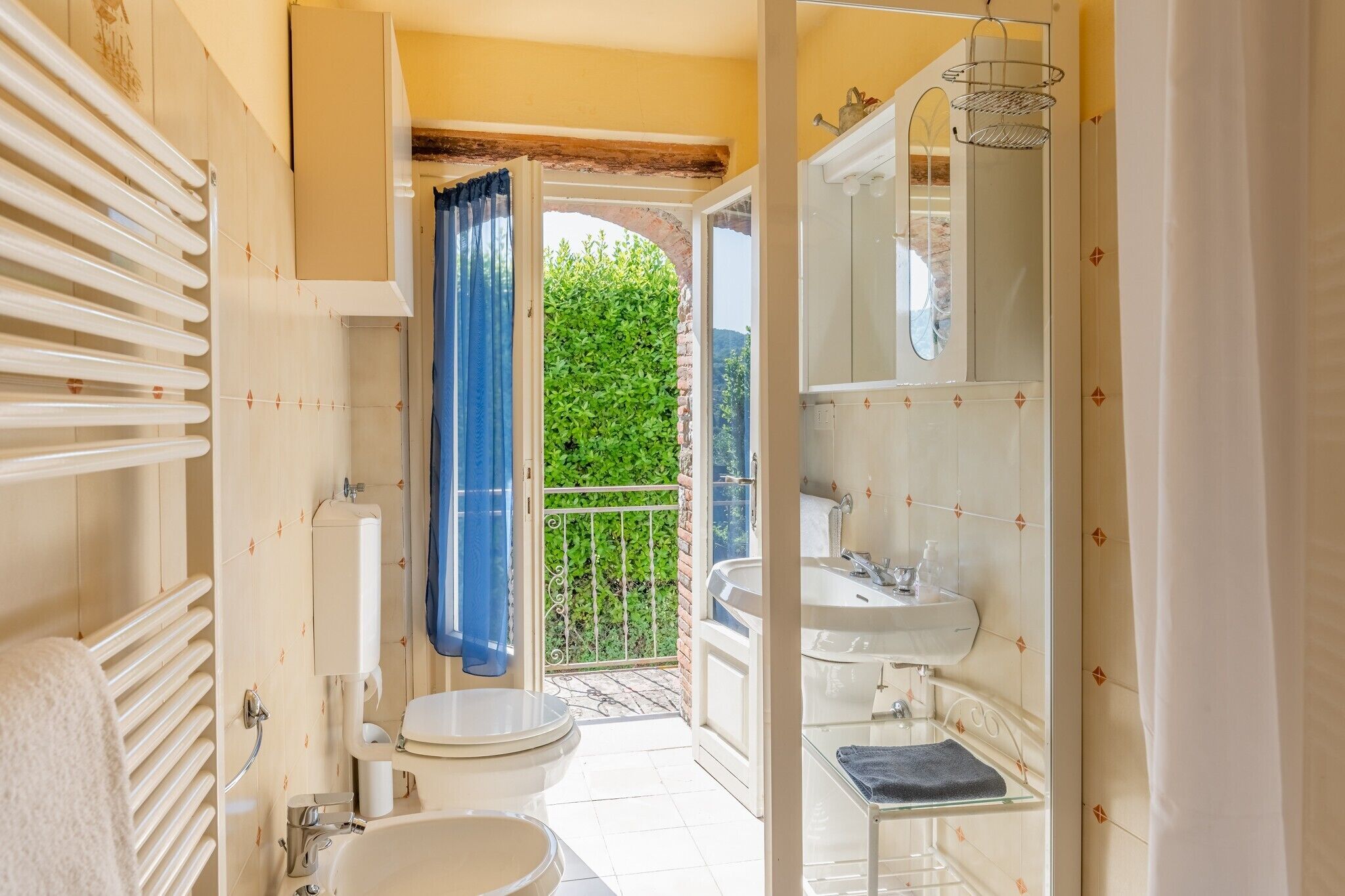 Baño con inodoro, lavamanos y zona de ducha, que da a un balcón con vistas a zonas verdes.