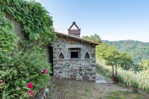 A stone building with a chimney, surrounded by greenery and a hilly landscape.