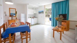 Una cocina moderna con armarios blancos, un comedor con mantel azul y una sala de estar con televisor y planta.