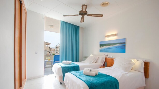 Habitación de hotel con dos camas, un ventilador de techo, vista al mar y un cuadro con una playa.