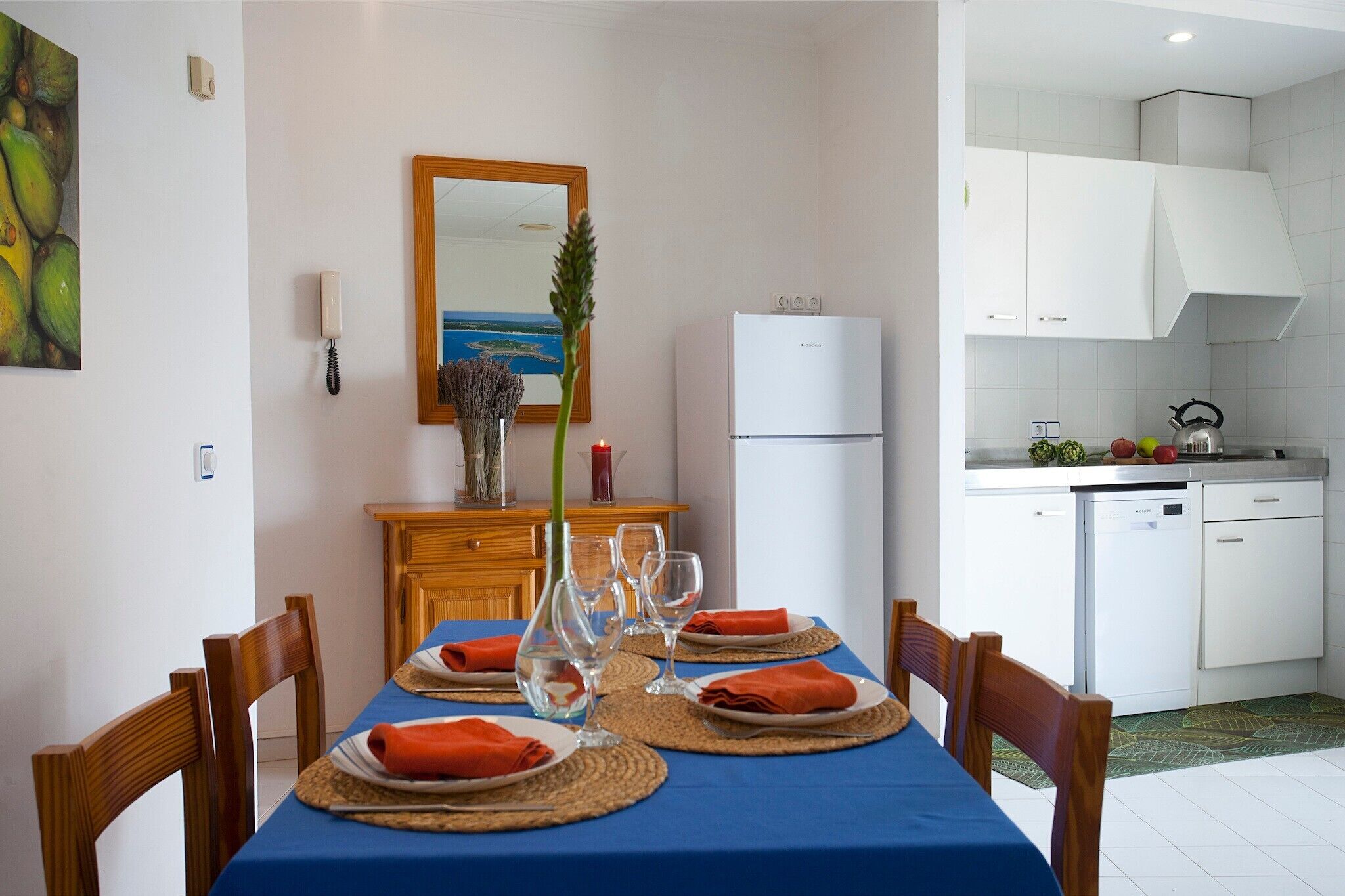 Un comedor con una mesa preparada para cuatro, una cocina con armarios blancos y un refrigerador.
