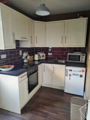 A compact kitchen with white cabinets, a black countertop, a microwave, a kettle, and a small refrigerator.