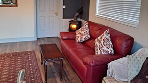 A living room with a red leather sofa, two patterned throw pillows, a wooden coffee table, a small rug, a fireplace, and a window with blinds. - 2-bed Apartment Sleeps 6 With Parking (Kildangan)