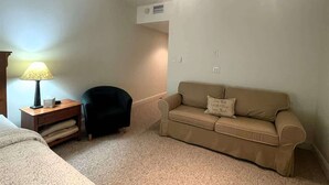 Een kleine, goed verlichte kamer met een bed, een nachtkastje met een lamp, een zwarte stoel en een beige bank met een kussen.