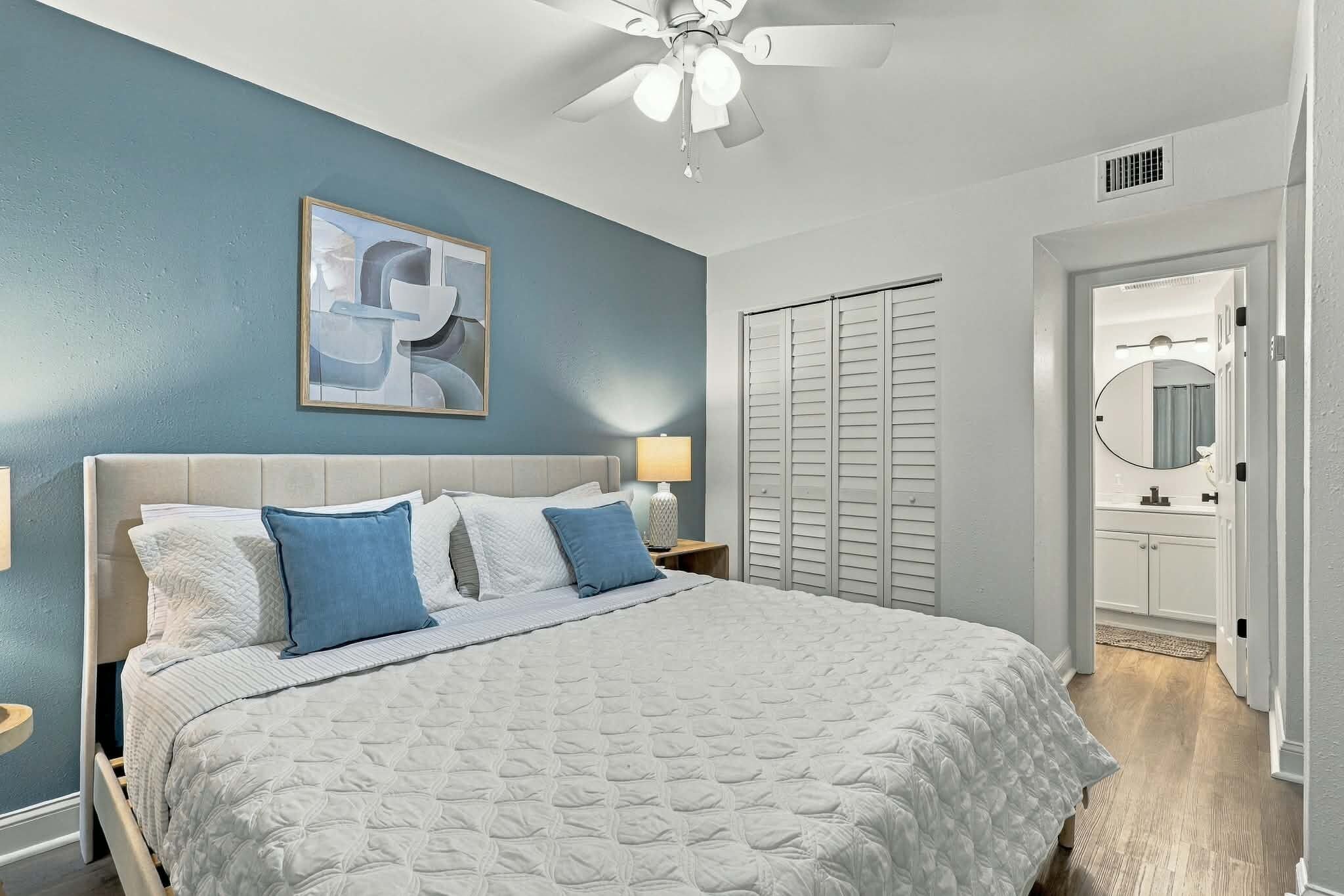 A bedroom with a bed, a ceiling fan, a nightstand, a lamp, a wardrobe, and a bathroom visible through an open door.