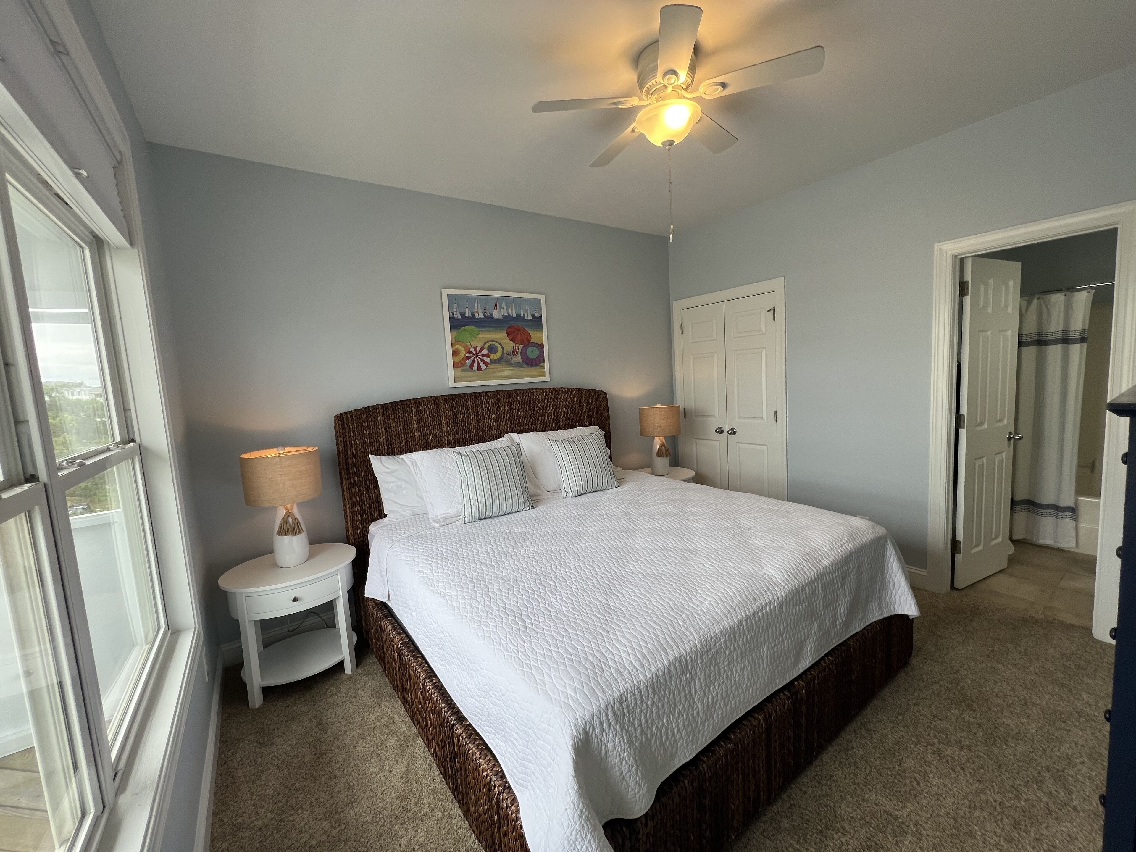 A bedroom with a bed, nightstands, a ceiling fan, a painting, and a door leading to a bathroom.
