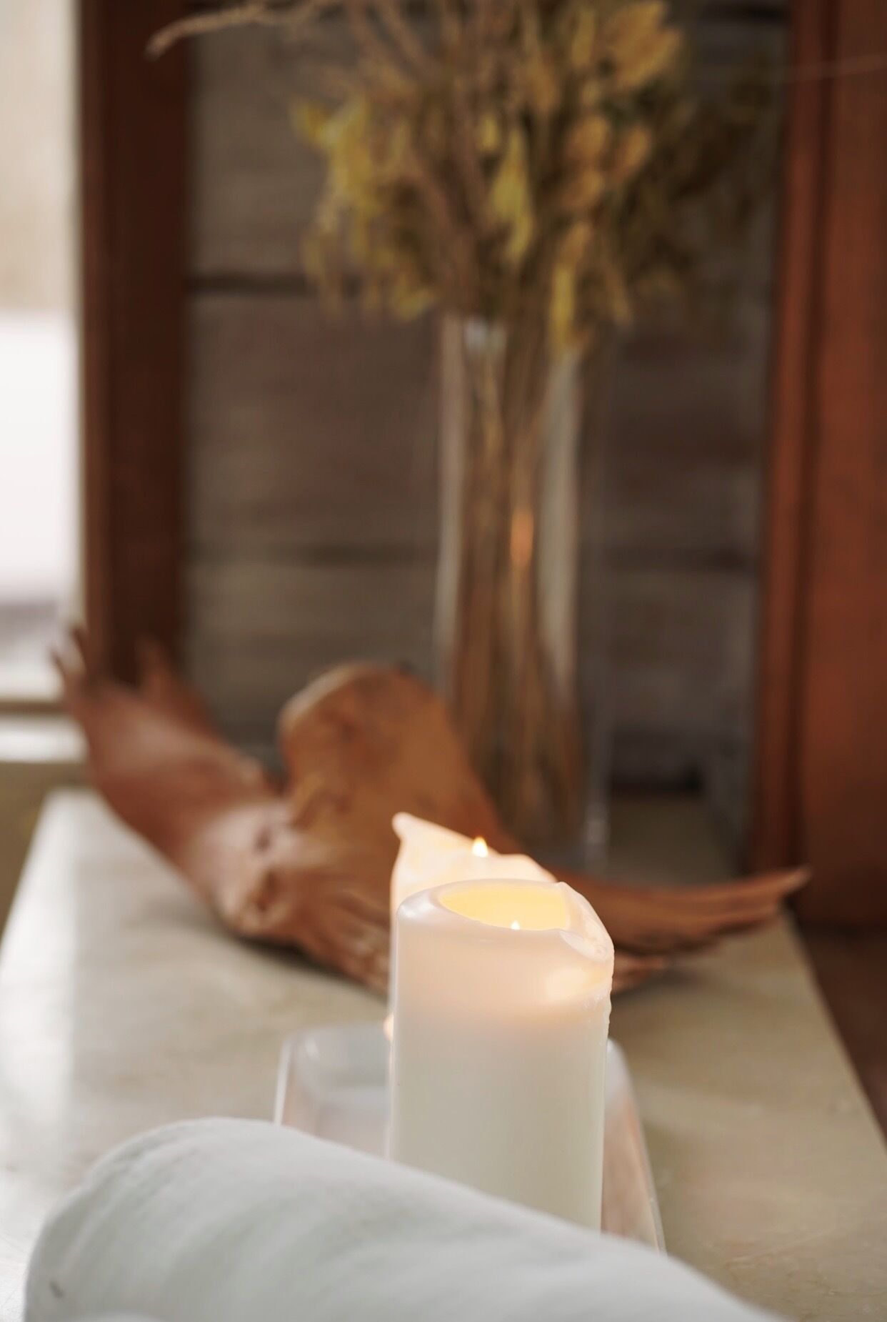 A candle on a table with a vase of dried flowers in the background.