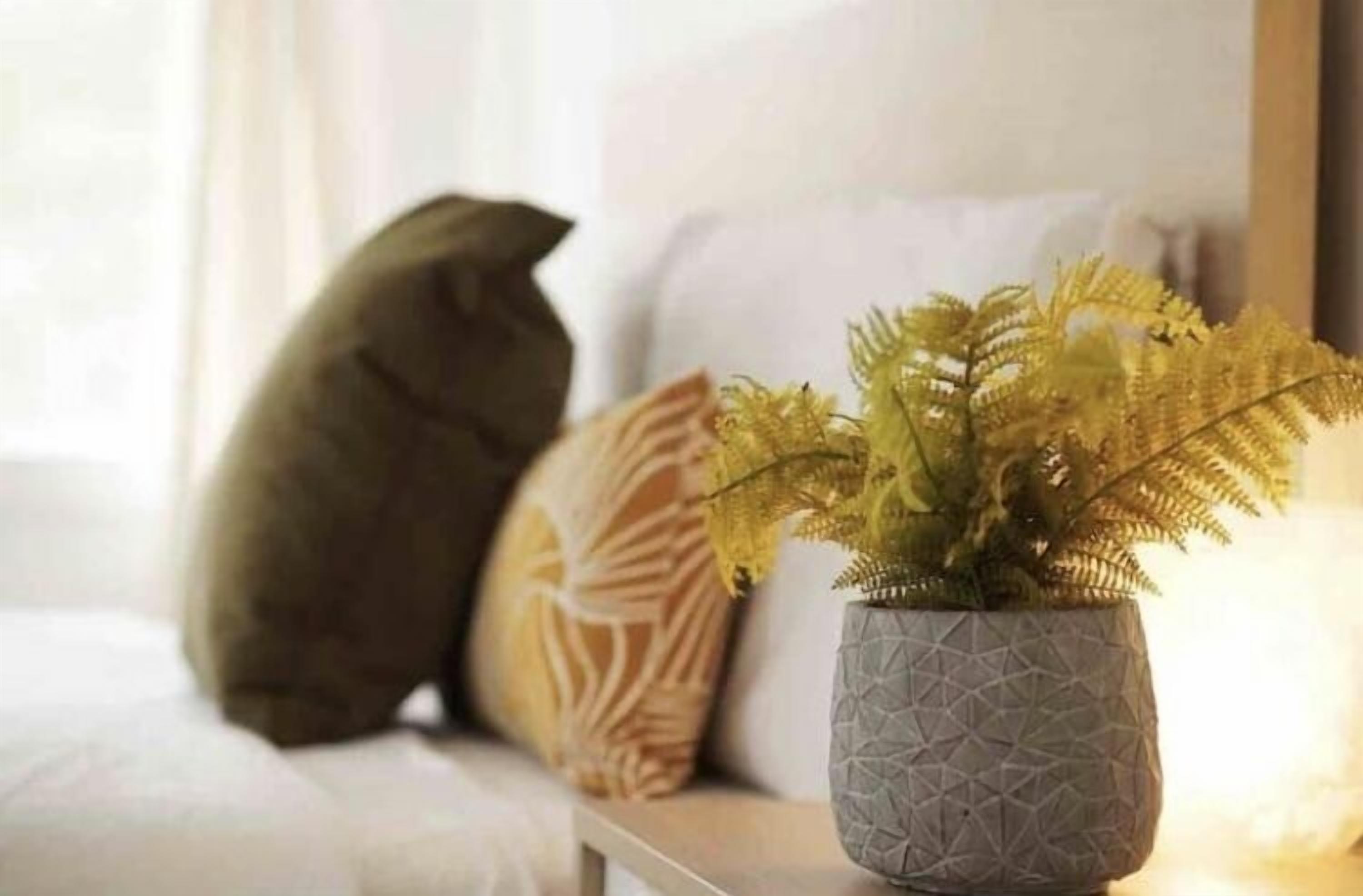 A cat sitting on a white couch with a textured grey vase containing a fern plant.