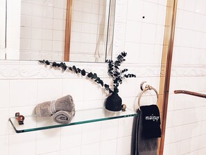 Une salle de bain avec une étagère en verre sur laquelle est posée une serviette enroulée, un vase noir avec une plante, et une serviette sur laquelle est écrit le mot « maquillage ».