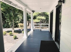Un porche couvert, doté de colonnes blanches et d’une porte noire, qui donne sur un jardin planté d’arbres et d’arbustes.