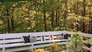 A wooden deck with a glass-top table surrounded by stools, white benches, and cushions. - The 212 Escape (Woodstock)
