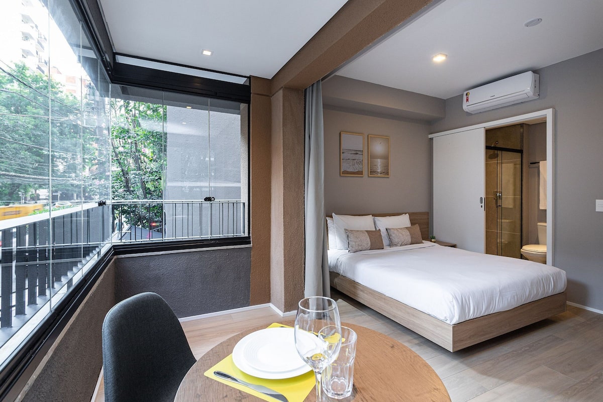 Une chambre d’hôtel avec un lit, une table à manger avec une chaise, un verre d’eau et une fenêtre donnant sur des arbres et des immeubles.