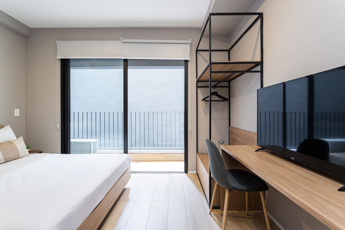 Ein modernes Hotelzimmer mit Bett, Schreibtisch, Sessel, Fernseher und einem Balkon mit Geländer.