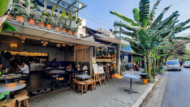 An outdoor cafe with tables and chairs, surrounded by potted plants and a building in the background.