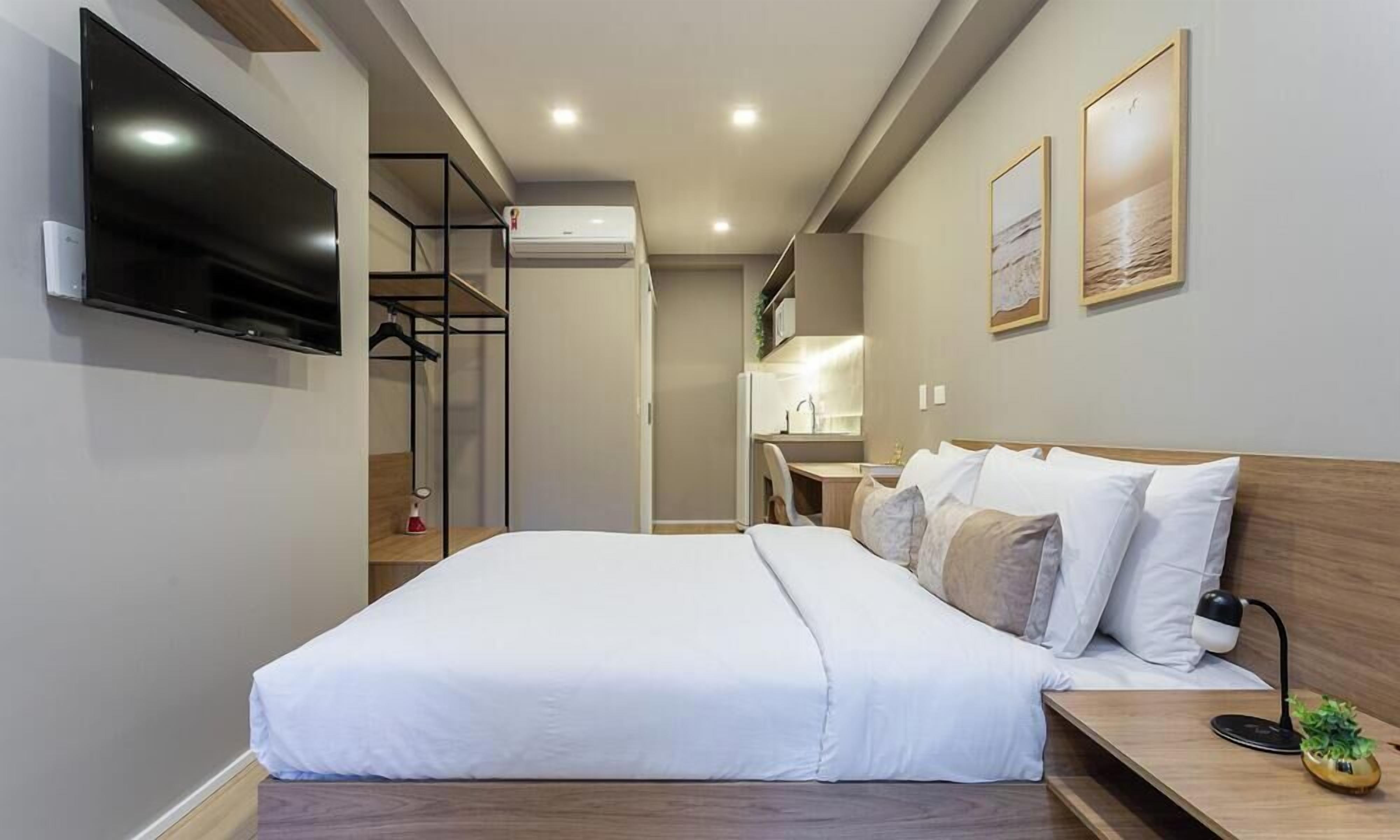 A modern hotel room with a large bed, a TV mounted on the wall, a small desk with a lamp, and a view of the kitchen area.