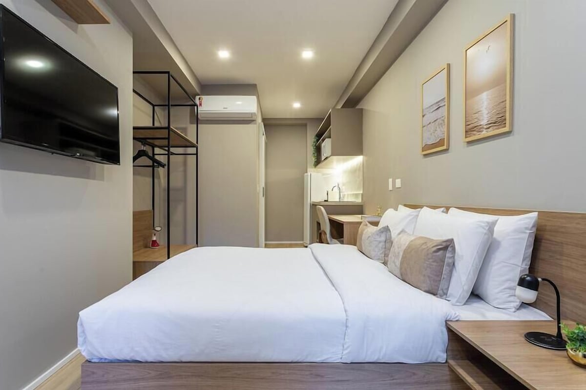 A modern hotel room with a large bed, a TV mounted on the wall, a small desk with a lamp, and a view of the kitchen area.