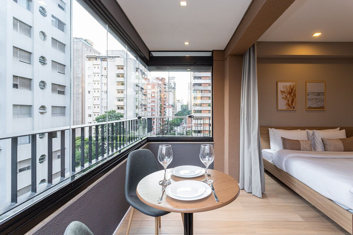 A modern hotel room with a balcony, a bed, a dining table with plates and glasses, and a view of high-rise buildings.