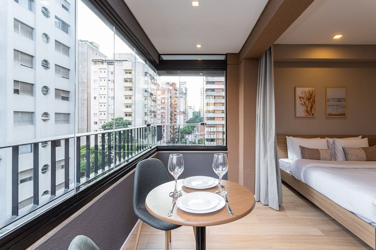Une chambre d’hôtel moderne avec un balcon, un lit, une table à manger avec des assiettes et des verres, et une vue sur des immeubles de grande hauteur.