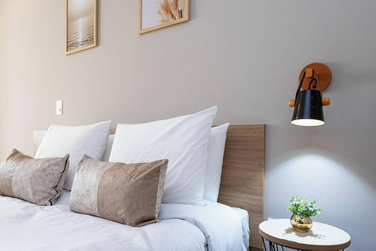 Un lit bien fait, avec des oreillers blancs et bruns, une table ronde ornée d’une plante et une lampe fixée au mur.