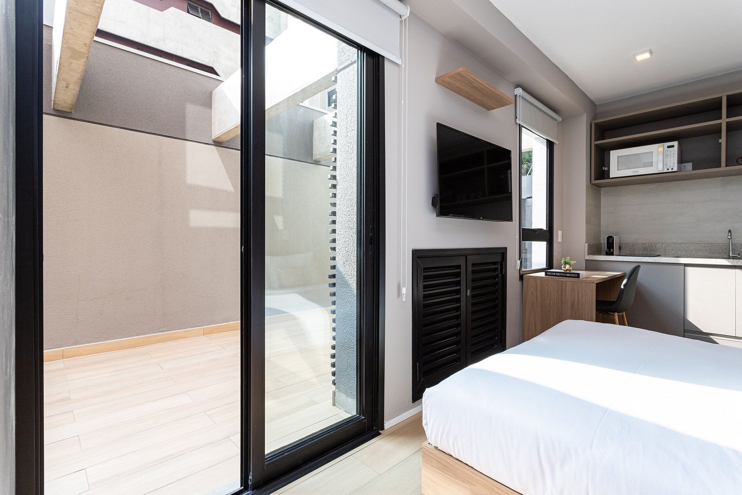 A modern bedroom with a large bed, a sliding glass door to a balcony, a built-in desk, and a mounted TV.