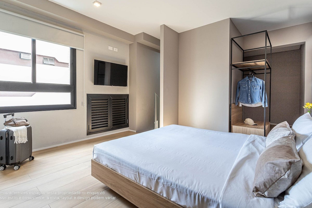 Une chambre à coucher avec un lit, un téléviseur fixé au mur, une fenêtre, une valise et une étagère suspendue avec des vêtements.