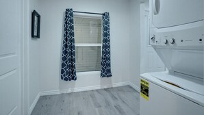 A laundry room with a washer and dryer, a window with patterned curtains, and a framed picture on the wall. - Step Into Paradise (River Oaks)