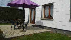 Eine Terrasse mit Tisch und Stühlen, einem violetten Sonnenschirm und einem weißes Gebäude mit einem Fenster.