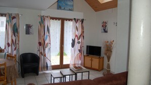 Ein Wohnzimmer mit Fernseher, einer Couch, einem Esstisch mit Stühlen und einer Schiebetür zu einem Außenbereich.