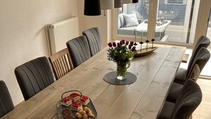 A dining room with a long wooden table, grey upholstered chairs, a vase with flowers, and a tray with pastries. - North Sea coast, charming Frisian house with fireplace, conservatory, sauna, beach chair (Wangerland)