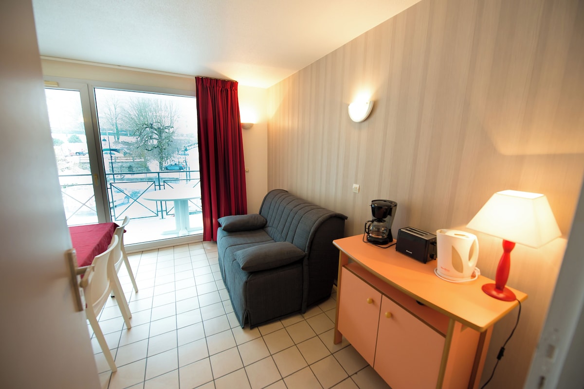 A compact room with a sofa, a small table with a lamp, a coffee maker, and a cabinet.