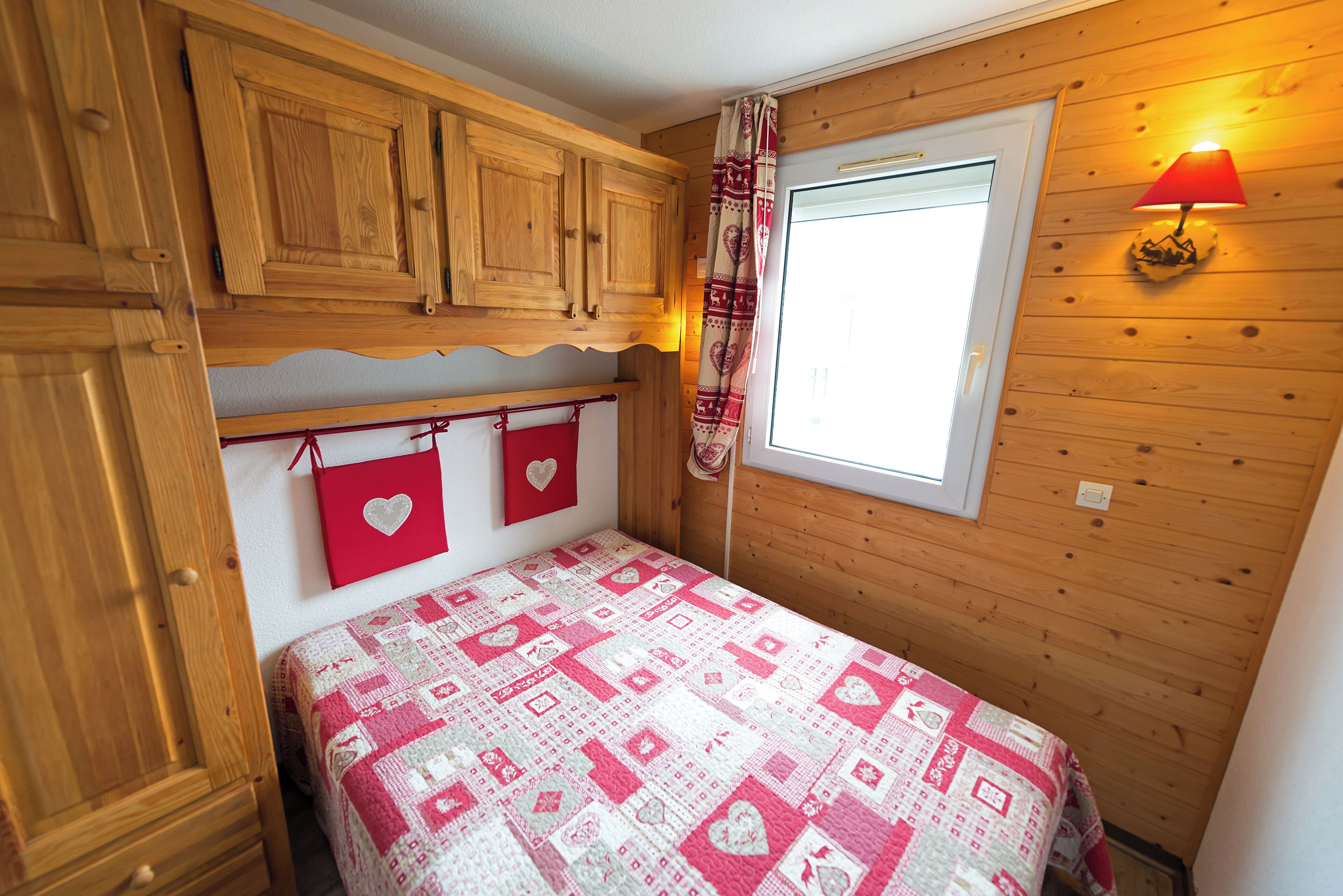 A bedroom with wooden paneling, a bed with a patterned quilt, a window with a red lamp, and a door with a heart-shaped decoration.