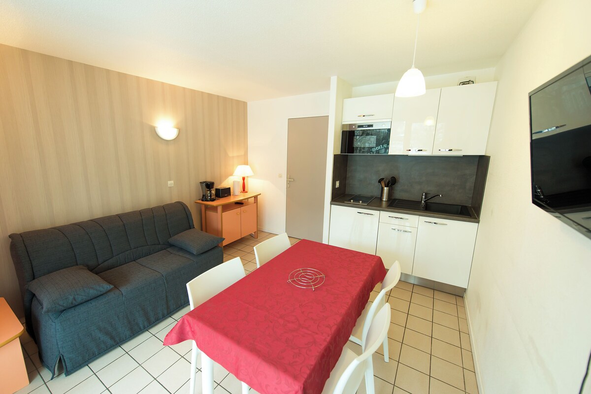 A compact living space with a sofa, dining table, kitchenette, and a TV mounted on the wall.