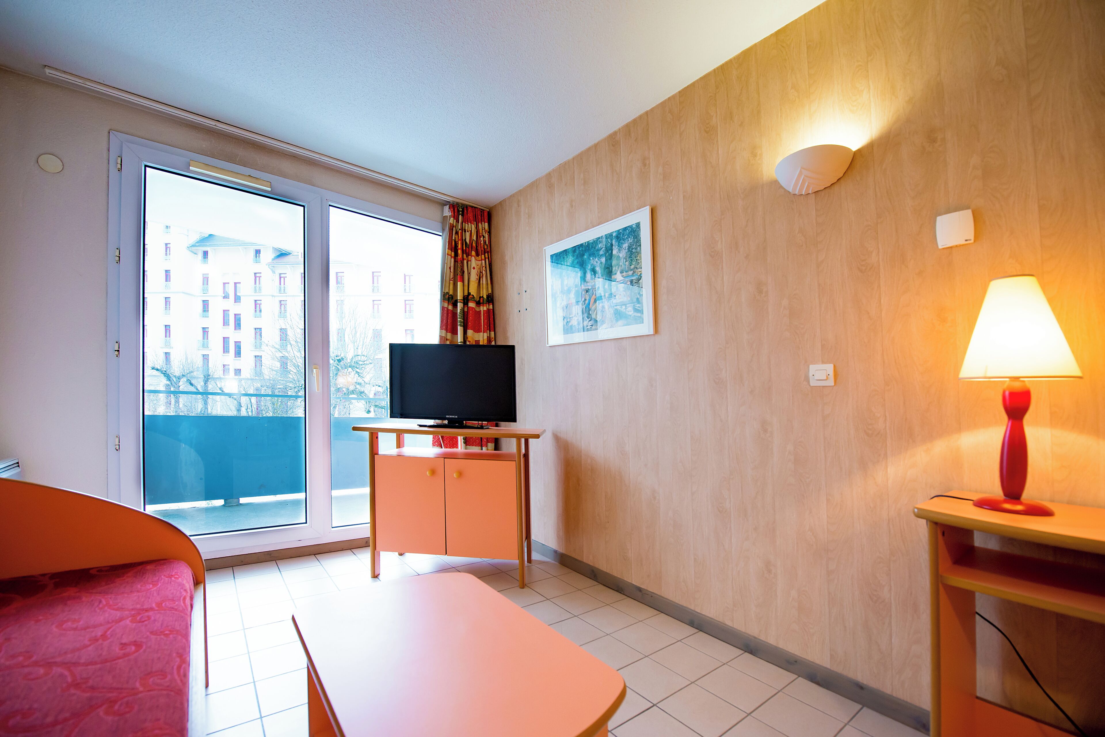 A hotel room with a bed, a desk, a television, a window with a view of buildings, and a wall-mounted lamp.