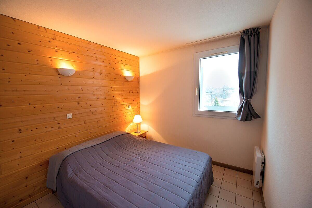 A room with a wooden wall, a bed with a grey bedspread, a window with curtains, and a wall-mounted heater.