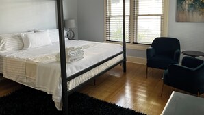 A four-poster bed with white linens, a lamp, a chair, and a table in a room with wooden flooring and a window with blinds. - 3 bedroom Craftsman bungalow in the heart of Lakeland (Lakeland)