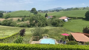 Ein Poolbereich mit Holzterrasse, umgeben von üppiger Vegetation und Blick auf sanfte Hügel und entfernte Häuser.
