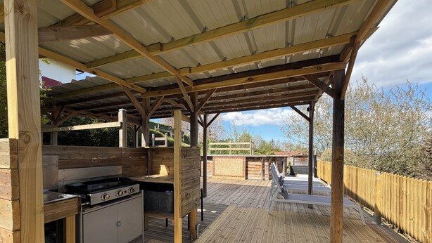 Eine Terrasse aus Holz mit Grill und Spüle, umgeben von einem Holzzaun und Bäumen.