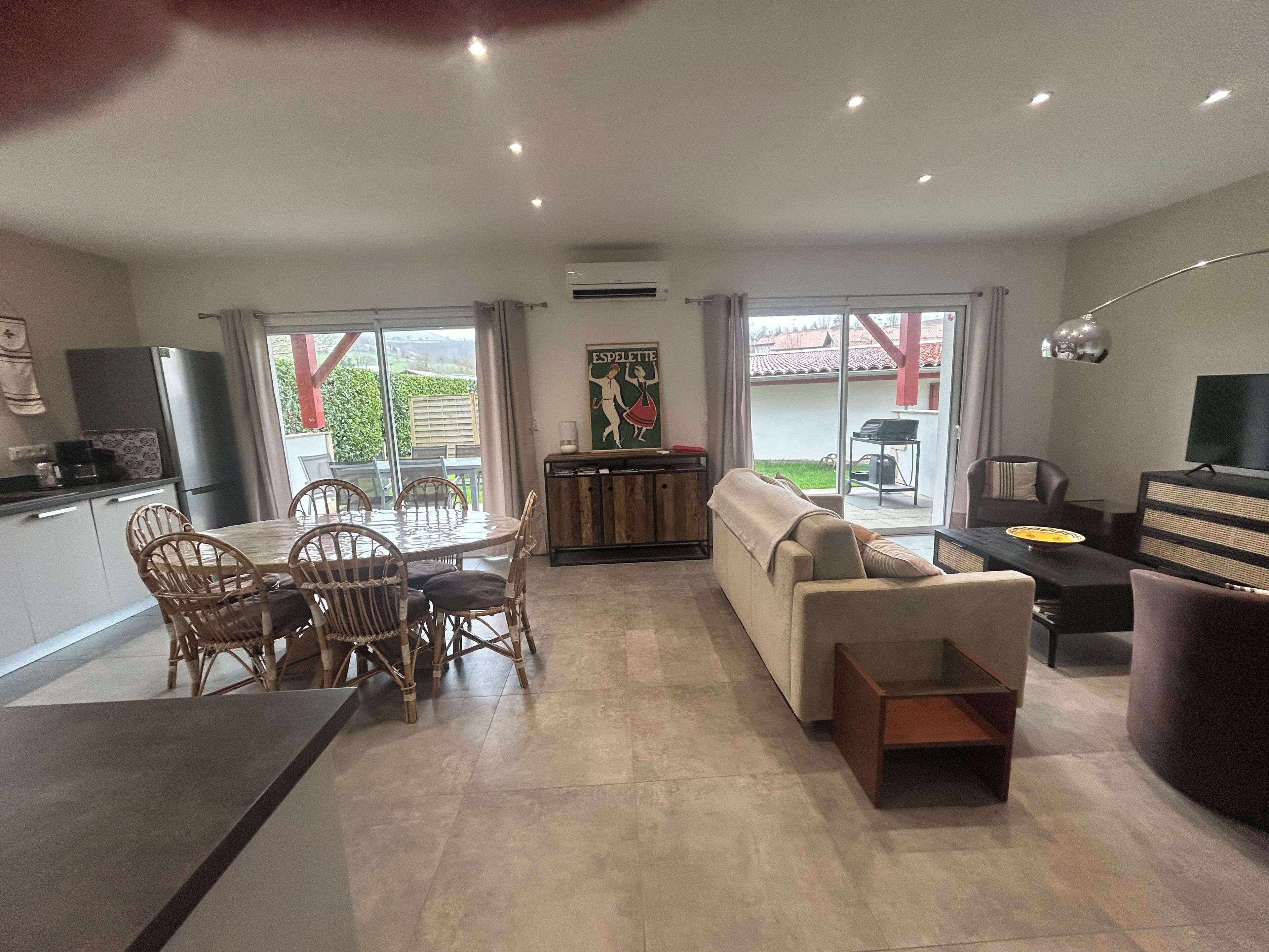 Una sala moderna con sofá, mesa de comedor y sillas. Hay una zona de cocina con refrigerador y mesa de comedor. La habitación tiene ventanas amplias y vistas al exterior.