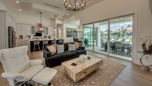 A spacious living area with a modern sofa, a white armchair, and a wooden coffee table. A large glass door leads to an outdoor patio with furniture.