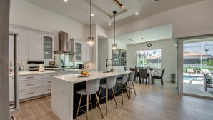 A modern kitchen with white cabinets, a central island with bar stools, and a dining area with a view of the outdoors.