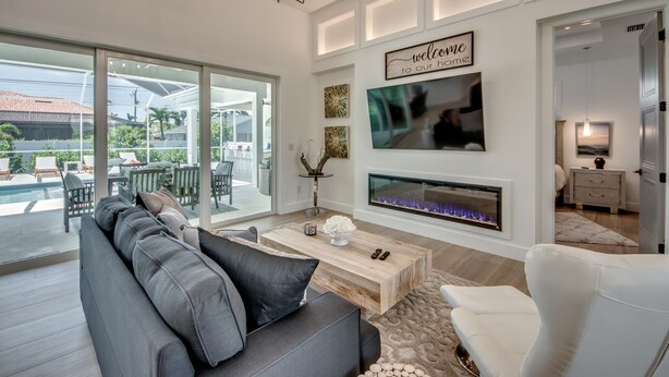 A modern living room with a gray sofa, a white armchair, a wooden coffee table, and a fireplace. There is a large window offering a view of a pool and outdoor furniture.