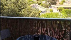 Uma varanda com uma mesa redonda e duas cadeiras, piso estampado, e vista para uma área residencial com árvores e edifícios.
