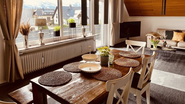 A modern living room with a dining area, large windows with curtains, a TV, and a sofa with cushions. - Charming Holiday House (Mastershausen)