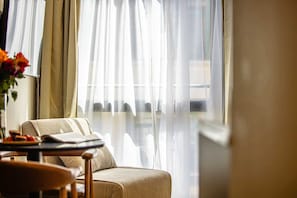 Huone, jossa on sänky, pöytä, jonka päällä on kirja, ja ikkuna, jossa on ohuet verhot.
